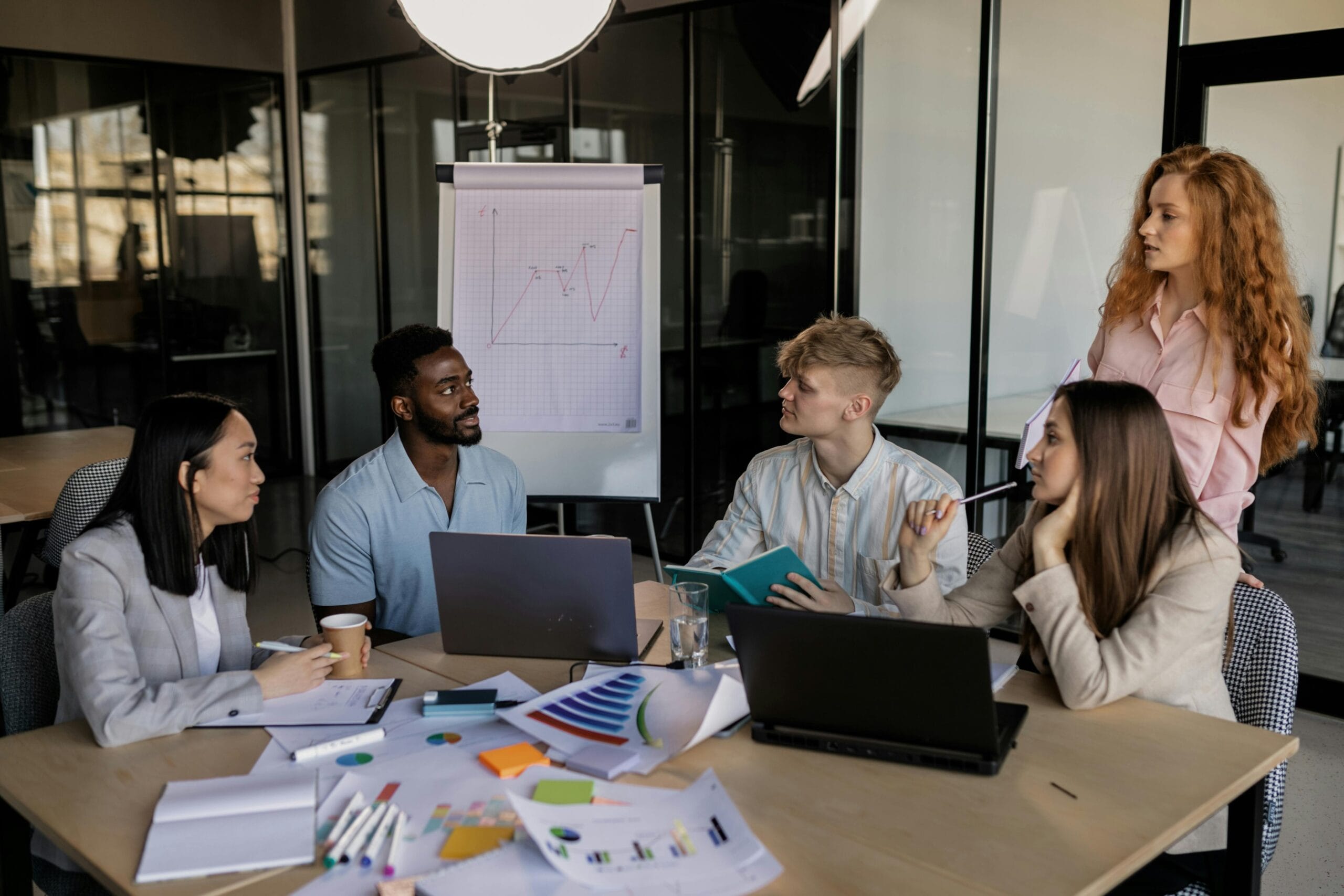 A team of four colleagues sitting at a table, actively discussing and collaborating, with one standing. Laptops are open, and a diagram is drawn on the board.