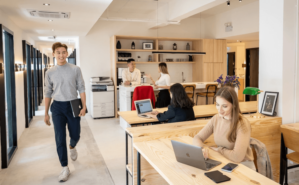 Modern coworking space with wooden desks, kitchen area, and people working or socializing casually