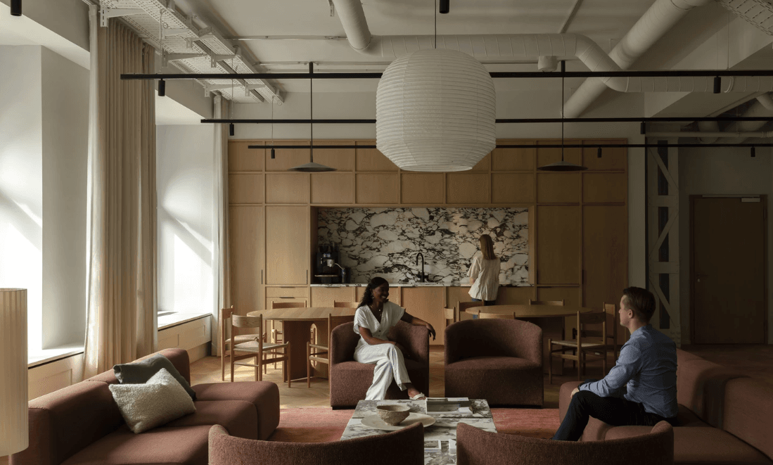 A stylish office lounge with a modern design, where two people are talking while another prepares a drink in the kitchen.