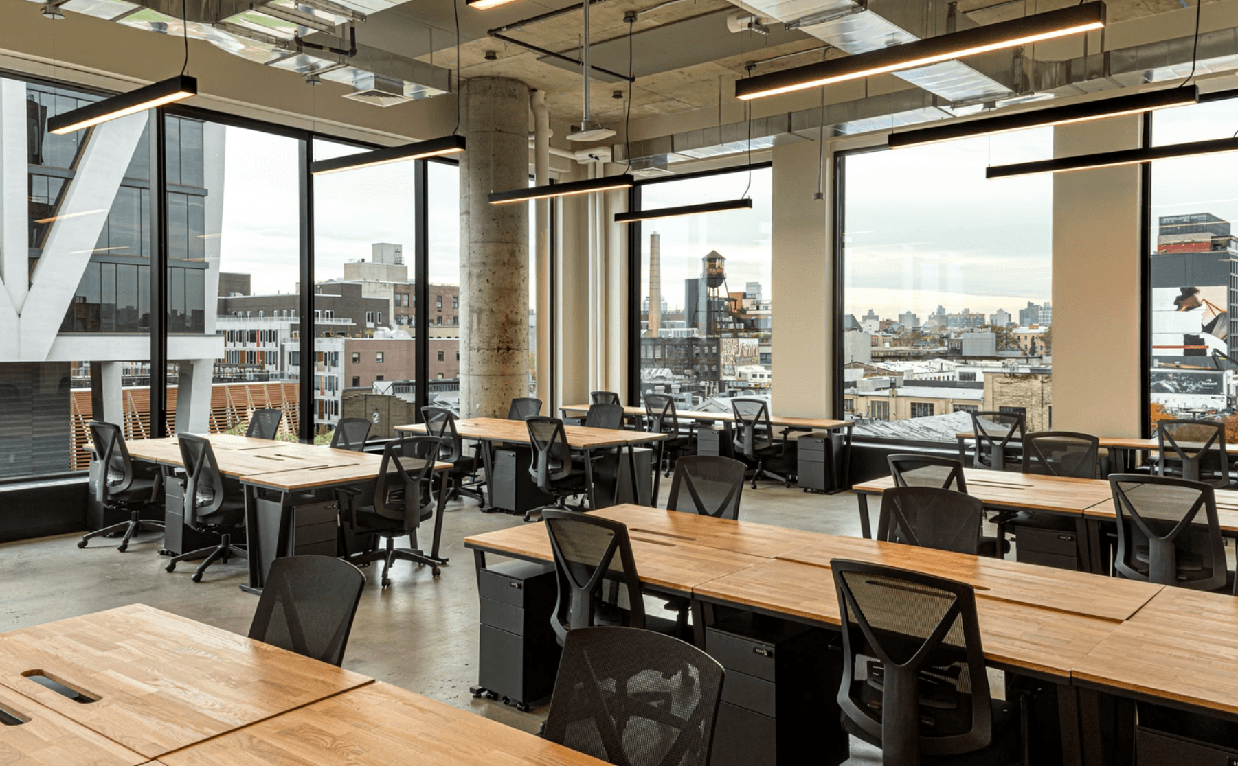 Spacious coworking office with wood desks, black chairs, and panoramic city views through large windows.