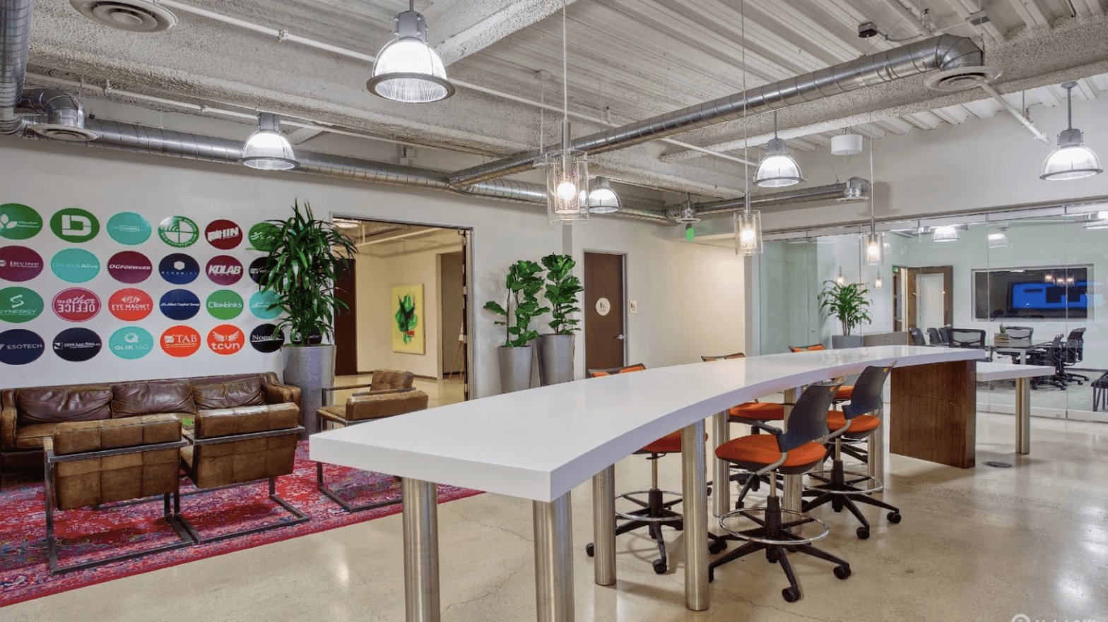 Modern coworking space with high tables, orange chairs, lounge area, wall logos, and indoor plants