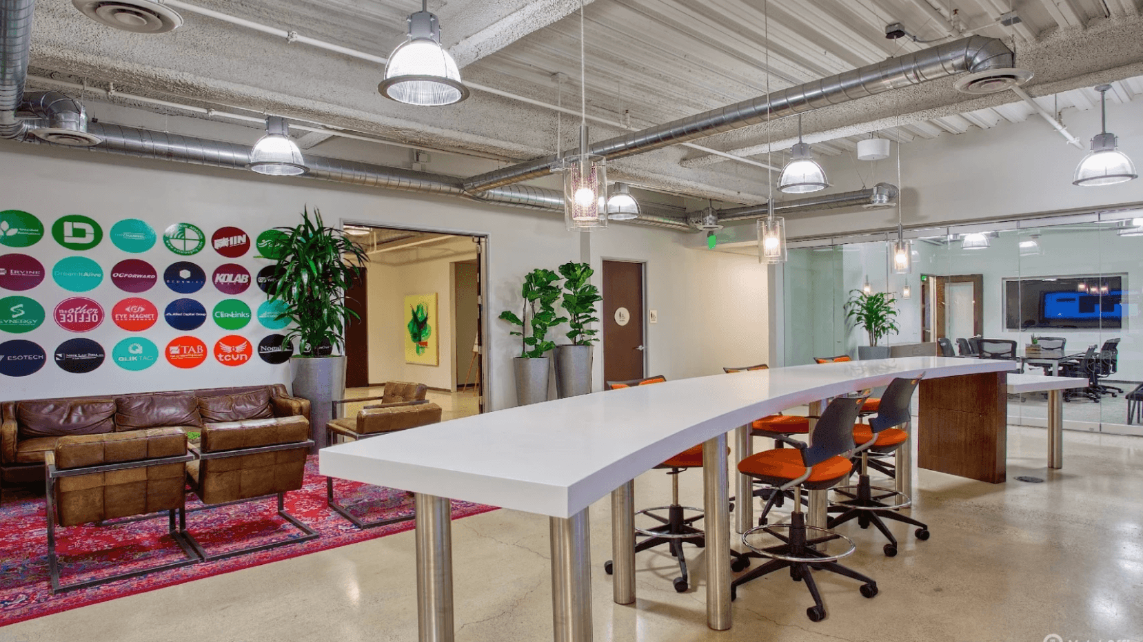Modern coworking space with high tables, orange chairs, lounge area, wall logos, and indoor plants